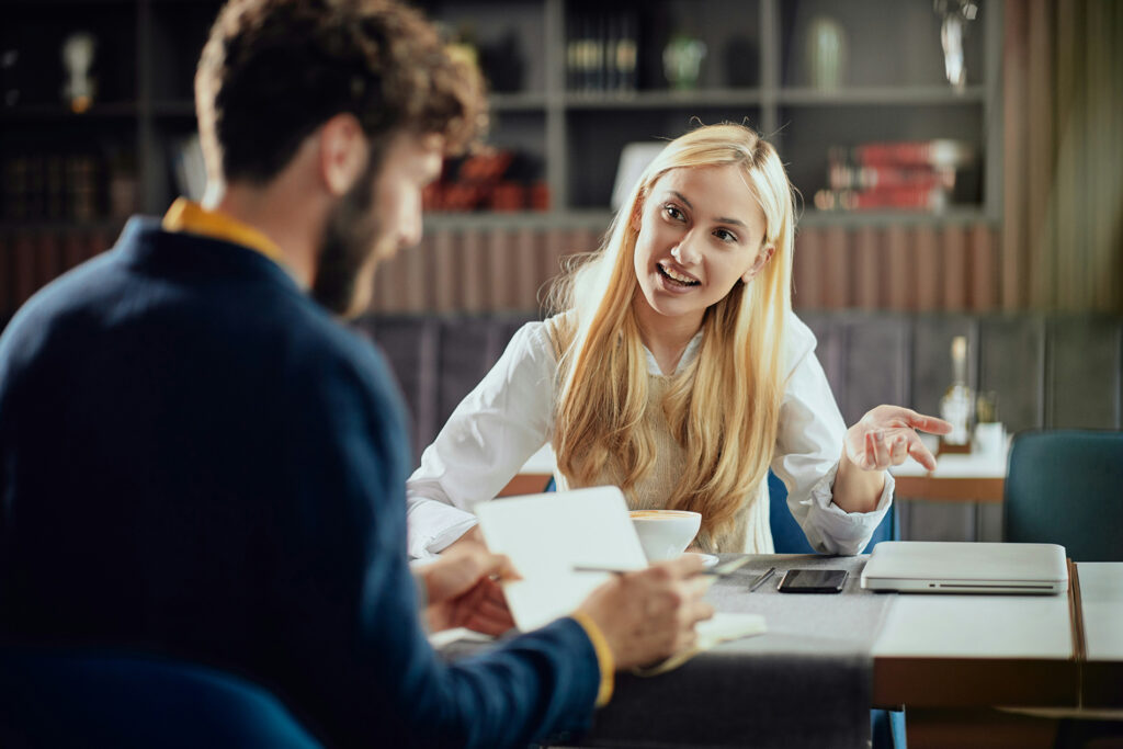Gestor anota enquanto colaboradora fala e gesticula durante um 1:1, com caderno, caneta e café na mesa