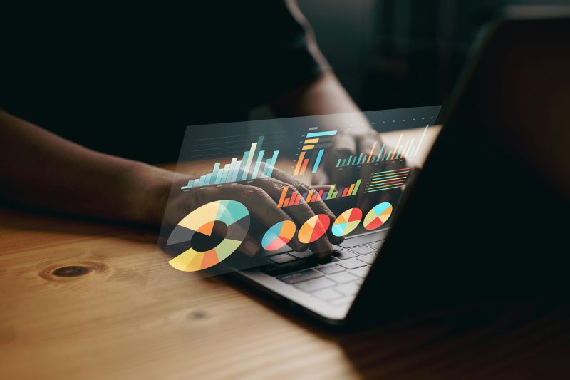 Mãos digitando em notebook com gráficos de dados sobrepostos, representando análise de dados no RH